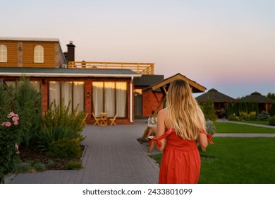 A girl with blond hair with her back to the camera walks into a country house at sunset