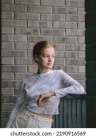 A Girl With Blond Hair Is Standing By The Railing. Medium Shot Portrait Against A Gray Brick Wall.