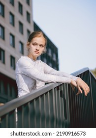 A Girl With Blond Hair Is Standing By The Railing. Medium Shot Portrait Against Sky And Building.