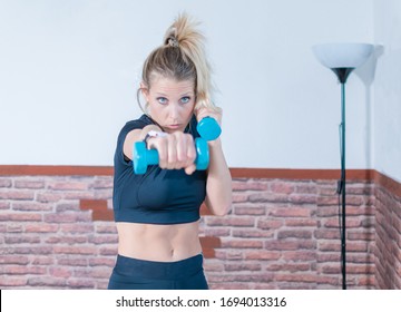 Girl, With Blonde Hair, Does Sports At Home In The Salon. Only Because Of The Insulation For COVID19. She Wears Sportswear And Trains By Boxing With The Shadow, And Punching With Blue Weights In His Ha