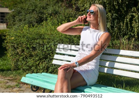 The girl the blonde in a white short dress, in sunglasses sits on a bench and speaks by phone in the park in the summer  