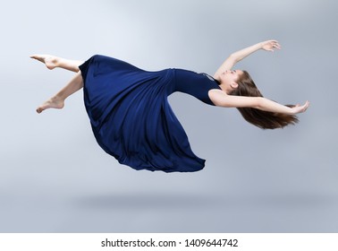 A Girl In A Blue Dress Is Floating In The Air. Dress And Hair Fluttering In The Wind. Flight. Fantasy. Insulation On Light Grey Background. Studio Photo