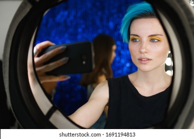 girl with blue hair and yellow eye makeup take a selfie on the phone in a beauty studio in front of a ring light on social media.