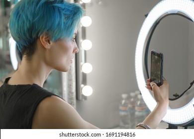 Girl With Blue Hair And Yellow Eye Makeup Take A Selfie On The Phone In A Beauty Studio In Front Of A Ring Light To Post To Social Media.