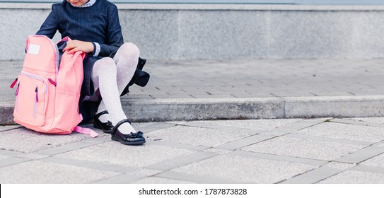 A Girl In A Blue School Dress, White Tights And Shoes Sits On The Steps With A Pink Backpack. Horizontal Photo. Copy Space