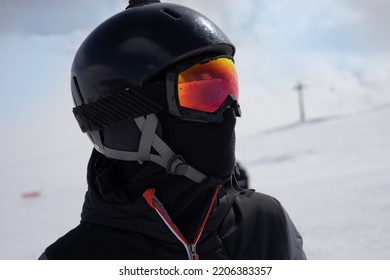 Girl Or Boy In Ski Helmet, Sunscreen Mask And Balaclava Close Up Stands Against The Backdrop Of Snow-covered Mountains And A Cloudy Sky. Winter. Sport And Travel Content  