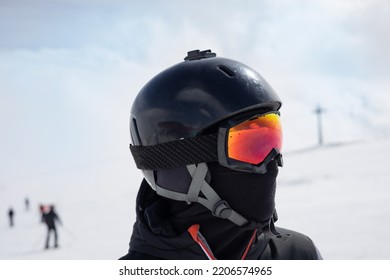 Girl Or Boy In Ski Helmet, Sunscreen Mask And Balaclava Close Up Stands Against The Backdrop Of Snow-covered Mountains And A Cloudy Sky. Winter. Sport And Travel Content  