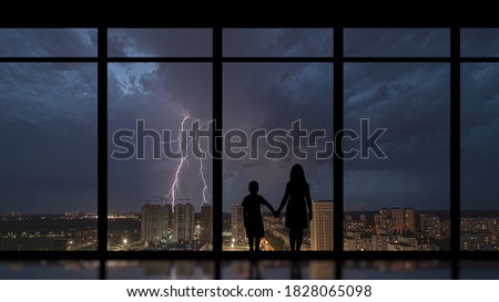 The girl and boy standing near a panoramic window against the night lightning