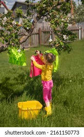 A girl in bright multicolored clothes and rubber boots hangs clean laundry on a clothespin under a tree in the garden. laundry detergent for washing colors children's clothes