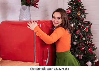 A Girl In A Bright Orange Blouse Hugs A Red Refrigerator Against The Backdrop Of A Christmas Tree. Gluttony Overeating On New Year's Eve.