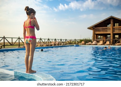 A Girl In Bright Pink Swimsuit And Black Swimming Goggles, Funny Jumping Into Deep Blue Pool With Clean Transparent Cool Water Against Background Of The Warm Summer Sunny Sunset