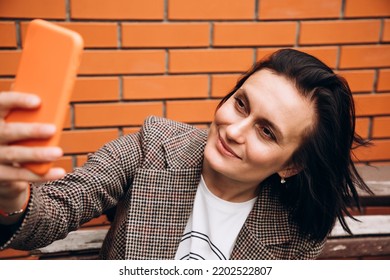 A girl in a brown jacket sits on a bench and takes a selfie. Portrait of a dark-haired girl against a background of a red brick wall - Powered by Shutterstock