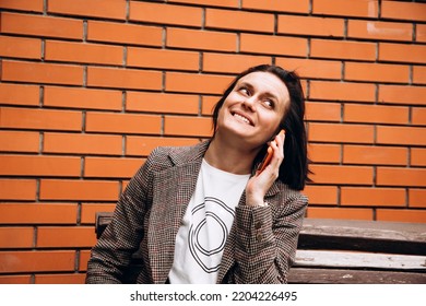 A girl in a brown jacket sits on a bench and talking on the phone, smiling. Portrait of a dark-haired girl against a background of a red brick wall, front view - Powered by Shutterstock
