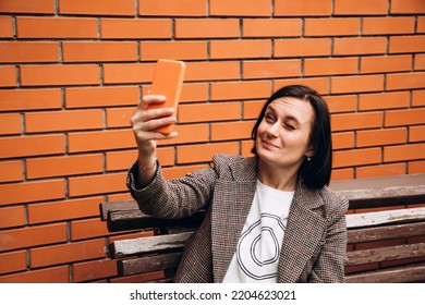 A girl in a brown jacket sits on a bench and takes a selfie with one eye closed. Portrait of a dark-haired girl against a background of a red brick wall - Powered by Shutterstock