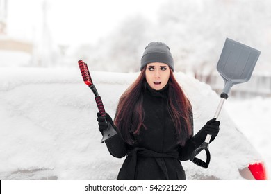 Girl With Brush And Shovel Removing Snow From The Car - Stressed Woman In Front Of A Frozen Auto In The Driveway
