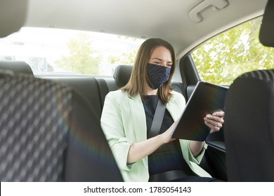 A Girl In Business Clothes Is Sitting In The Back Seat Of A Car With A Protective Mask On Her Face And Holding A Tablet. Work On The Way To The Office