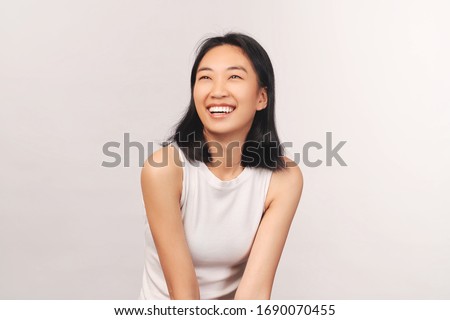 Girl cackles and looks up at empty space for text and your ad. Beautiful young woman Asian appearance with black hair brown eyes stands isolated white background in Studio