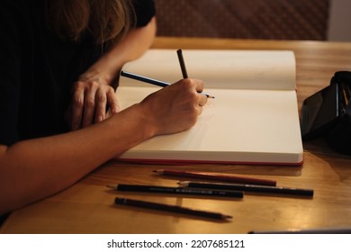 A Girl In A Cafe Draws In A Sketchbook With Colored Pencils. Notepad, Pencils, Black Pencil Case.