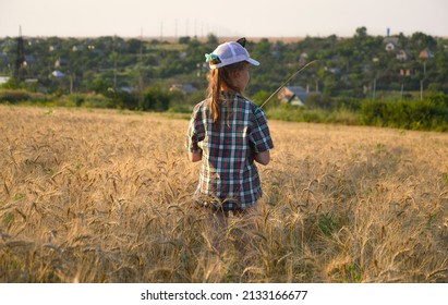 Girl In A Cap And A Country-style Shirt In A Ripened Wheat Field At Sunset. Summer Evening Walk Outdoors