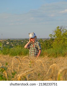 Girl In A Cap And A Country-style Shirt In A Ripened Wheat Field At Sunset. Summer Evening Walk Outdoors