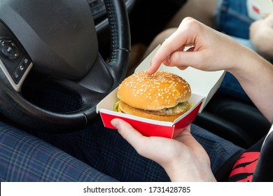 The Girl In The Car Holds A Burger In Her Hands. Fast Food, Takeaway, Snacks On The Road.