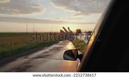Girl in car window waves her hand and catches wind at sunset, Wet road after rain. Happy young woman driver rides car, Hand catches glare of sun. Girl student, rides in car, Dreamy look, carefree ride