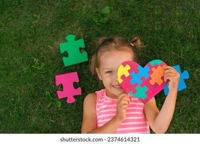 A girl with card made of colored puzzle pieces is lying on the grass, demonstrating support for people with autism spectrum syndrome