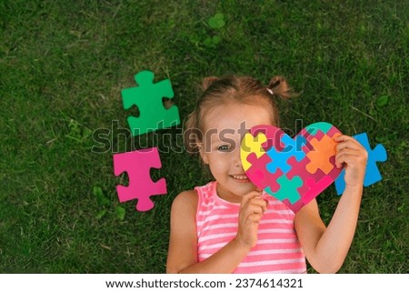 A girl with card made of colored puzzle pieces is lying on the grass, demonstrating support for people with autism spectrum syndrome