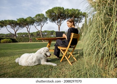Girl caressing Maremmano-Abruzzese Sheepdog and using laptop computer at wooden table on green mediterranean lawn. Concept of weekend, rest and remote work. Idea of friendship of human and pet
