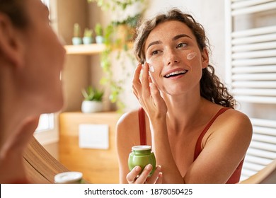 Girl Caring Of Her Beautiful Skin Face Standing Near Mirror In The Bathroom. Young Woman Applying Moisturizing Cream On Face. Smiling Natural Girl Holding Little Green Jar Of Ecological Cosmetic Cream