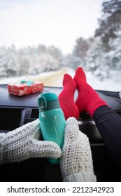 Girl Carry Gift Sits In Red Socks And Knitted Mittens In A Car In Winter And Warms Herself With Coffee From A Thermos, Against The Background Of Snow Winter Forest. Travel, Trip And Christmas Concept