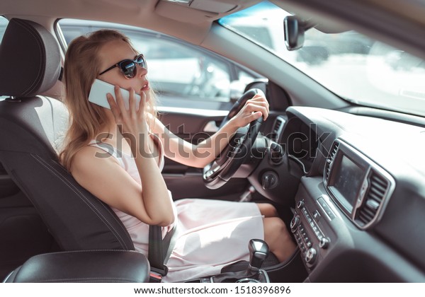 girl in car\'s interior calls on phone, looks in\
rearview mirror, parks at parking lot of shopping center, holds it\
fastened by steering wheel. Woman in summer sunglasses in city\
wearing pink dress.