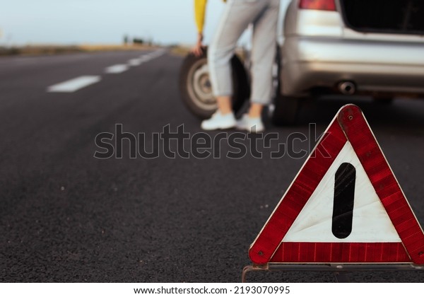a\
girl in casual clothes stands near the rear wheel of the car and\
rolls the spare wheel to change. Car breakdown, the wheel has been\
pierced. Close-up and focus on the emergency stop\
sign,