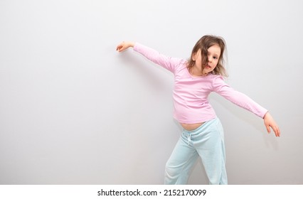 Girl, Caucasian 6 Years Old, In A Pink Coat On A Gray Background. The Child Is Happy And Smiling, Waving His Arms And Dancing