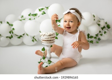 A Girl Celebrates Her First Birthday And Eats A Birthday Cake. Decorations For A Child's Birthday Made Of Balloons And Ivy. A Cheerful Happy Child On The Day Of His Birth.