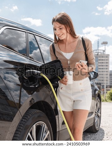 Girl charging her electric car at the parking lot near the city business center with skyscrapers visible in the background