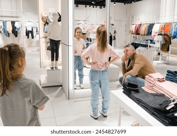 A girl checks her reflection in a fitting room mirror with an adult sitting nearby. - Powered by Shutterstock