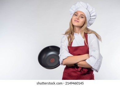Chica con sombrero de chef. El chef dobla los brazos y mira la cámara. Rubia con uniforme de chef. Una chica con una sartén frita sobre un fondo claro. Recetas de cocina. Secretos culinarios. Entrenamiento de cocina.