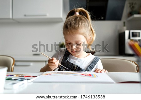 A girl child of 4 to 7 years old sits in the house and paints on paper. The child draws a rainbow, focused on the picture. Large woman portrait.