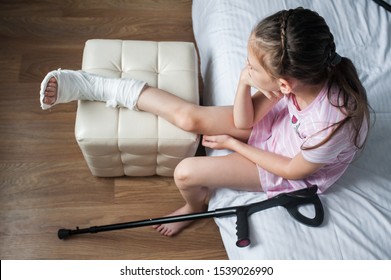 Girl Child With A Broken Leg In A Cast Is Sitting On A Bed With Crutches.