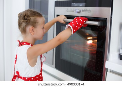 Girl Child In Gloves Waiting For Baking Muffins Or Cupcakes Near Oven.The Little Girl Looking At Tasty Fruitcake In The Oven. Cooking Process On Kitchen