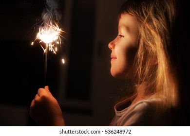 Girl, Child Holds In His Hands A Sparkler On A Dark Background At Home