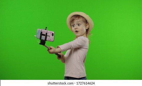 Girl Child Makes Selfie On Mobile Phone Using Selfie Stick. Kid With Pink Hair In Hat And Pink Blouse Using Smartphone Social Network App For Video Call, Blogging. Green Screen At Studio. Chroma Key