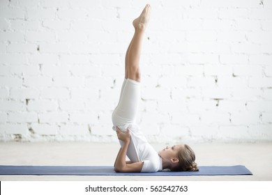 Girl child practicing yoga, standing in salamba sarvangasana exercise, supported Shoulder stand pose, working out wearing sportswear, t-shirt, pants, indoor full length, white loft studio background  - Powered by Shutterstock