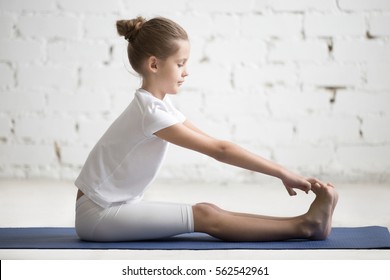 Girl Child Practicing Yoga, Stretching In Paschimottanasana Exercise, Seated Forward Bend Pose, Working Out, Wearing Sportswear, Indoor Full Length, White Loft Studio Background 