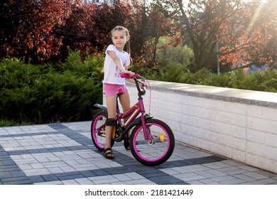 Girl Child Rides In The Park On A Red Bike And Is Dressed In Summer Clothes. A Cute Child With Pigtails Sits On A Bike At Sunset. The Child Is Happy With The Ride On A Beautiful Bike