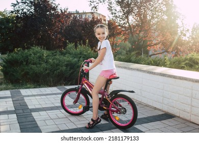 Girl Child Rides A Red Bike And Is Dressed In Summer Clothes. Happy Smiling Child With Pigtails Sits On A Bicycle At Sunset In The Park. The Child Is Happy With A Ride On A Beautiful Bike