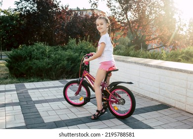 Girl Child Rides A Red Bike And Is Dressed In Summer Clothes. Happy Smiling Child With Pigtails Sits On A Bicycle At Sunset In The Park. The Child Is Happy With A Ride On A Beautiful Bike