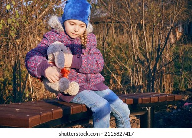 A girl child sits on a park bench and hugs a plush bunny. A place for walking and relaxing.
