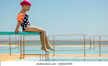 Girl Child Sitting On A Diving Platform At The Swimming Pool. Girl In Swim Gear Sitting On Diving Spring Board.